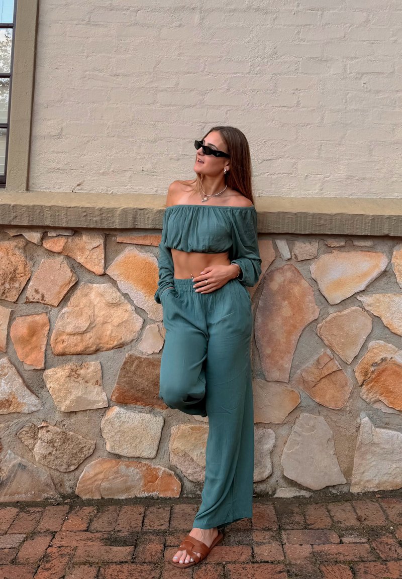 Γυναίκα με τιρκουάζ μπλούζα off-shoulder και παντελόνι, καφέ σανδάλια, μαύρα γυαλιά ηλίου, στέκεται μπροστά σε πέτρινο τοίχο με το ένα πόδι λυγισμένο.
