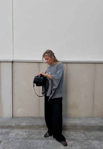 Suéter gris de gran tamaño, pantalones negros de pierna ancha y zapatos de malla negros. Bolso negro con correa corta que se sostiene mientras se mira dentro. Fondo claro.