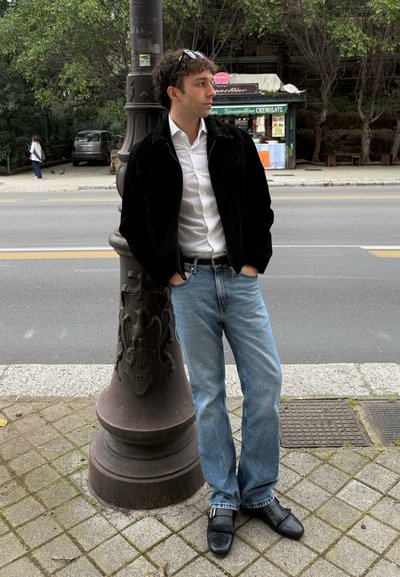 Joven con chaqueta negra y vaqueros, apoyado en un farol decorado en una acera cerca de una calle y un quiosco.