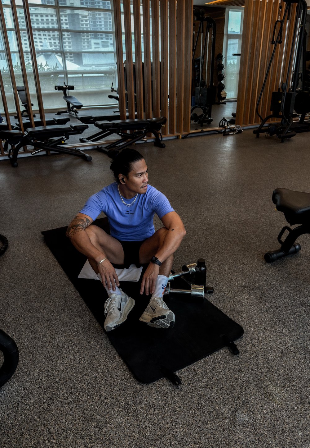 Atletisk mand i en lilla skjorte og sorte shorts sidder på en sort måtte i et fitnesscenter, omgivet af vægtudstyr og store vinduer.