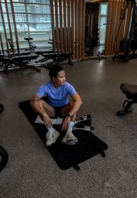 Atletisk mand i en lilla skjorte og sorte shorts sidder på en sort måtte i et fitnesscenter, omgivet af vægtudstyr og store vinduer.