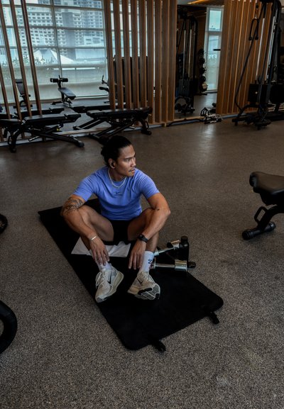 Hombre atlético con camisa morada y pantalones cortos negros sentado sobre una colchoneta negra en un gimnasio, rodeado de equipo de pesas y grandes ventanales.