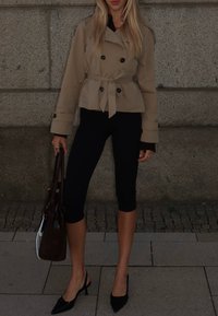 Trench coat beige con bottoni doppi, vita cinturata, abbinato a leggings neri e tacchi neri a punta, mentre tiene una borsa marrone.
