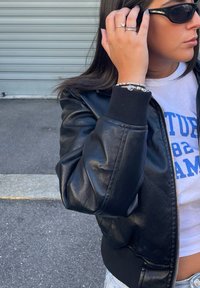 Giubbotto bomber in pelle nera con polsini a coste, indossato sopra una maglietta grigia. Gli accessori includono più braccialetti e anelli, e occhiali da sole scuri.