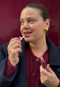 Läppglans i ett klart rör med en silverapplicator hålls av en person. De bär en djup bordeauxfärgad texturerad tröja och en marinblå kappa.