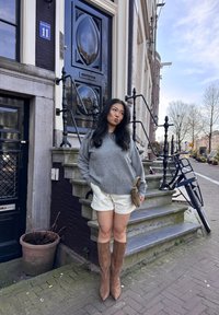 Grå genser, stripete hvite shorts og brune knelange støvler. Står på steintrappen, med en svart dør og en sykkel i bakgrunnen.