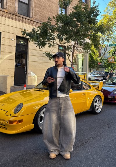 Hombre con vaqueros grises de gran tamaño, chaqueta de cuero negra y gorra negra, de pie en la calle sosteniendo un teléfono cerca de un coche deportivo amarillo.