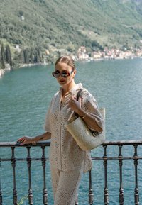 Outfit di pizzo color beige chiaro con maniche corte; porta una borsa a tracolla in paglia intrecciata; indossa occhiali da sole oversize con motivo tartaruga.