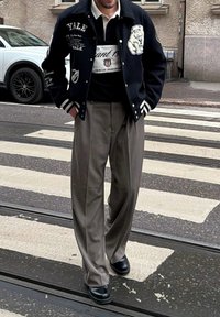 Veste varsity bleu marine foncé avec des bandes blanches et le logo Yale, associée à un pantalon large gris clair et des chaussures noires, traversant une rue.