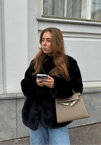 Μαύρο παλτό από τεχνητή γούνα, με φαρδιές μανίκες, συνδυασμένο με μια μπεζ δομημένη τσάντα και ανοιχτόχρωμα jeans. Κρατάει ένα smartphone, στέκεται έξω.