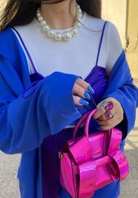 Roupas em camadas roxas e azuis, colar de pérolas oversized, bolsa vibrante rosa com laços e unhas azuis ousadas com anéis coloridos.