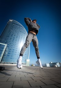 Sportists pelēkā flīsa džemperī ar kapuci un pelēkās sporta biksēs lec uz bruģētas virsmas, valkājot baltas sporta kurpes. Fonā redzama moderna stikla ēka.