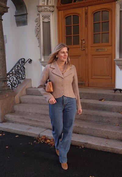 Chaqueta ajustada de cuadros en tonos marrones, suéter de cuello alto en marrón claro, jeans de talle alto azules, botas de tacón en color marrón claro y bolso pequeño marrón, caminando por escalones de piedra.