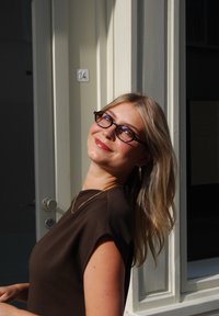Braunes kurzärmliges Oberteil, goldene Halskette, runde schwarze Brille, lange blonde Haare, vom Sonnenlicht beleuchtet vor einer hellen Wand mit Türgriff.