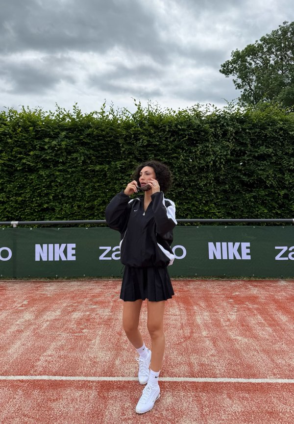 Fekete sportfelszerelésű nő napszemüveget igazít egy vörös salakos teniszpályán, háttérben zöld sövénnyel és Nike molinókkal.