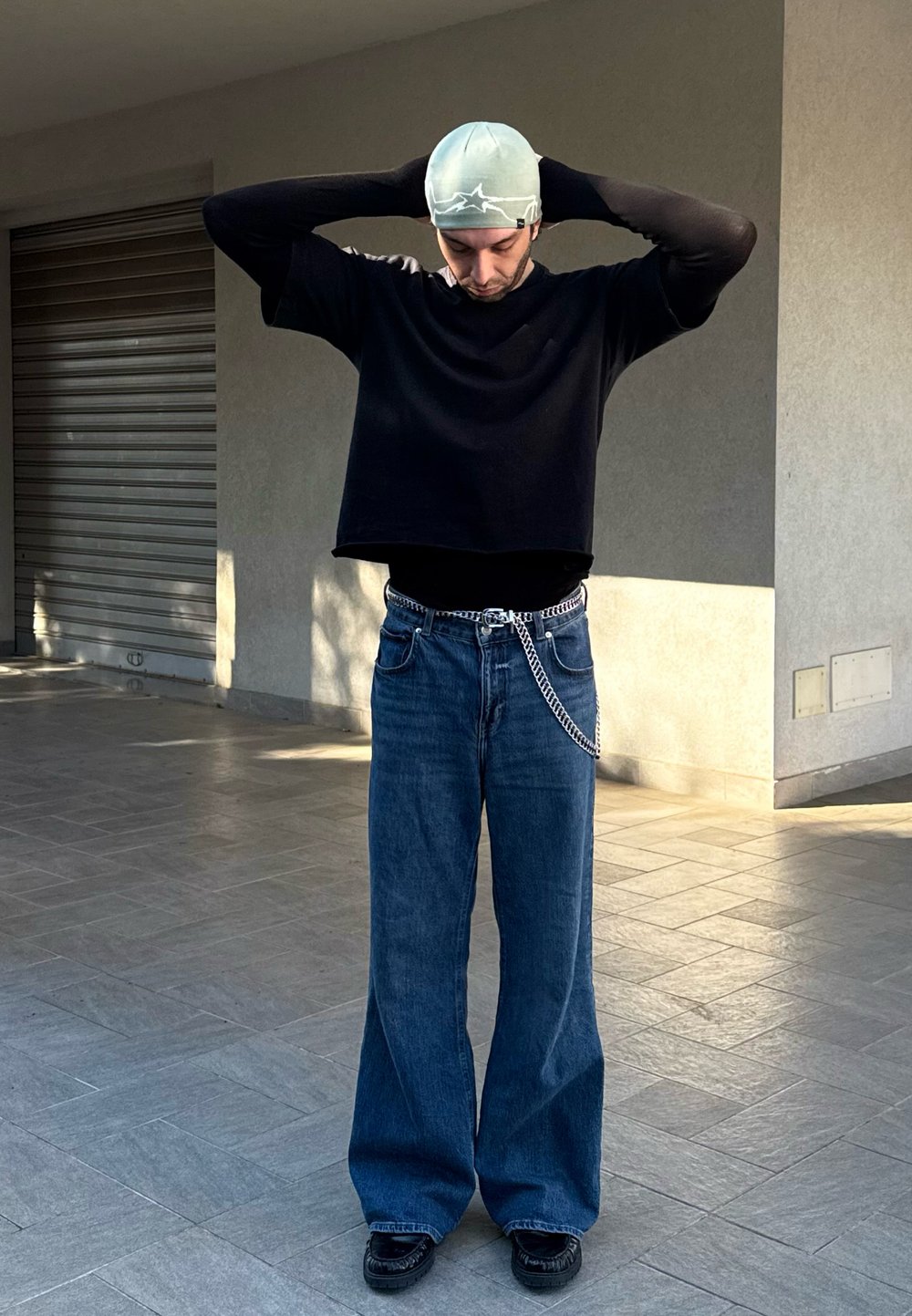 Μαύρη κοντή μακριά μπλούζα με μανίκια, φαρδιές μπλε τζιν παντελόνες με ασημένια αλυσίδα στη ζώνη και μαύρα παπούτσια. Το φόντο διαθέτει μια υφασμάτινη τοίχο.