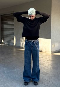 Μαύρη κοντή μακριά μπλούζα με μανίκια, φαρδιές μπλε τζιν παντελόνες με ασημένια αλυσίδα στη ζώνη και μαύρα παπούτσια. Το φόντο διαθέτει μια υφασμάτινη τοίχο.