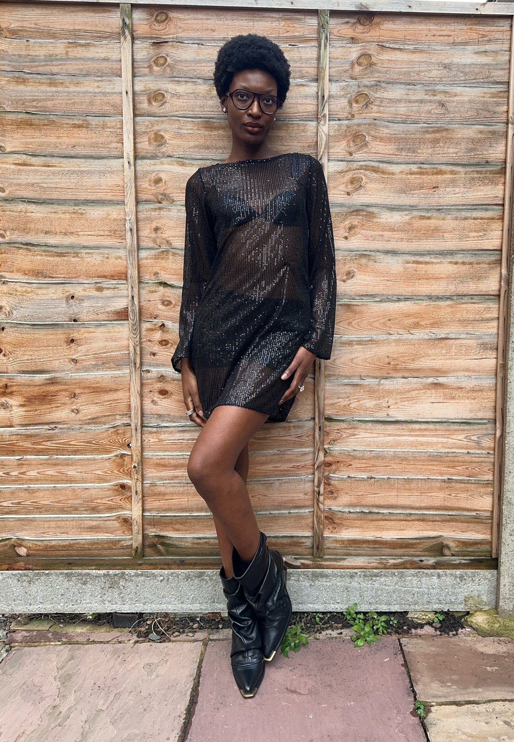 Juoda mini suknelė su blizgučiais, ilgomis rankovėmis, permatoma medžiaga ir aptempta siluetu. Derinama su juodais laisvo silueto kulkšnies aulinukais. Medienos fonas.