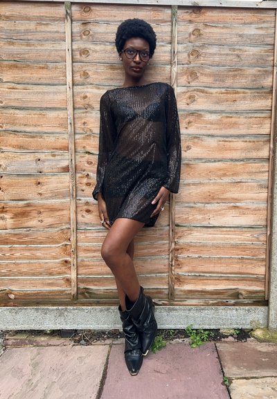 Vestido mini negro con lentejuelas, mangas largas, tela transparente y silueta ajustada. Combinado con botines negros holgados hasta el tobillo. Fondo de madera.