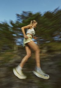 Persoană tânără în mișcare, purtând un maiou alb și pantaloni scurți galbeni deschiși, asortați cu adidași albi și șosete de sport gri.