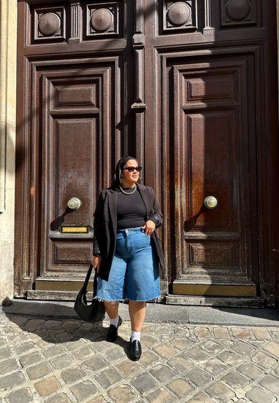 Blazer negro sobre una blusa negra, pantalones cortos de mezclilla azul claro con dobladillo deshilachado, zapatos negros, llevando un bolso negro, frente a una puerta de madera.