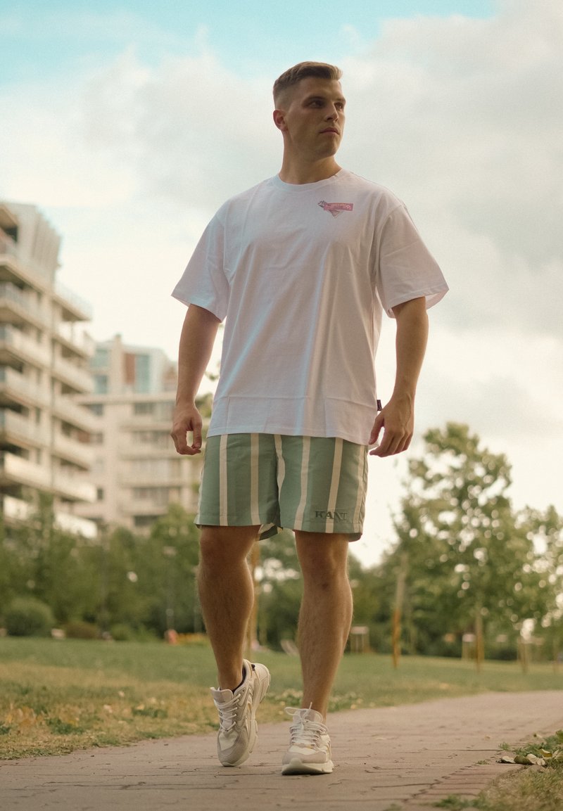 Homme en t-shirt blanc et short rayé vert marchant sur un chemin pavé dans un parc avec des bâtiments et des arbres en arrière-plan.