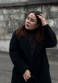 Svart ullkappa med breda slag, ovanpå en mörk tröja. Långa, vågiga bruna lockar och en silverring. En enkel texturerad bakgrund.