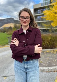 Burgundi színű, galléros, cropped kabát ezüst gombokkal, amelyet magas derekú világoskék farmernadrággal viselnek. A haj hullámos, a háttérben őszi lombozat látható.