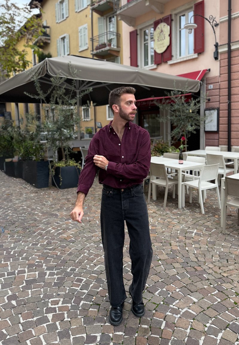 Camicia marrone chiaro con bottoni, jeans neri a gamba larga e scarpe nere; in piedi su pavé, ambientazione da caffè all'aperto con tavoli e vegetazione.