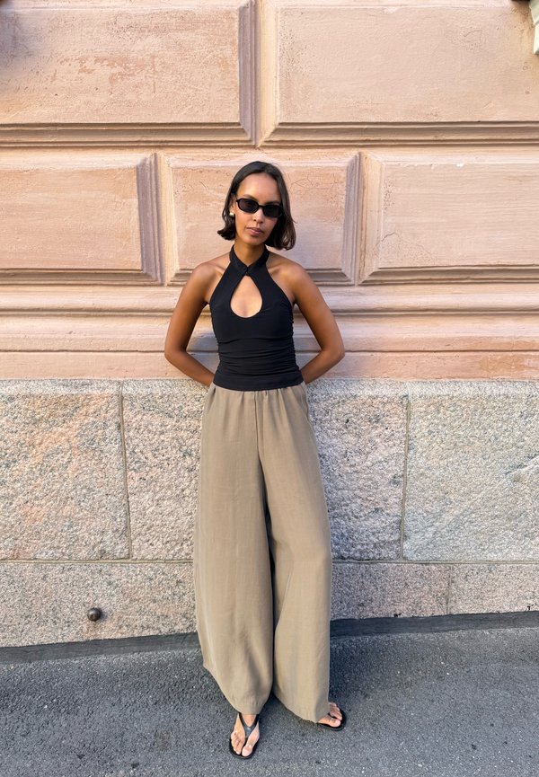 Mujer que lleva top halter negro, pantalones beige de pierna ancha, gafas de sol y sandalias, de pie contra una pared texturizada de piedra y yeso.