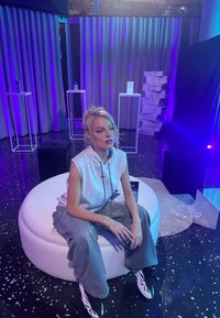 Jeune femme aux cheveux blonds attachés, portant un sweat à capuche sans manches et un pantalon ample, assise sur un pouf rond blanc dans une pièce faiblement éclairée avec un éclairage violet.