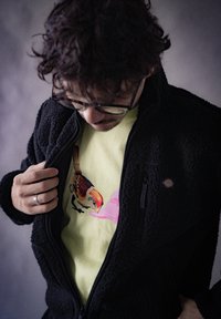 Giacca in pile nera aperta che rivela una maglietta gialla chiara con una grafica di un uccello e un'illustrazione di un cappello rosa. Tessuti lisci e texturizzati.