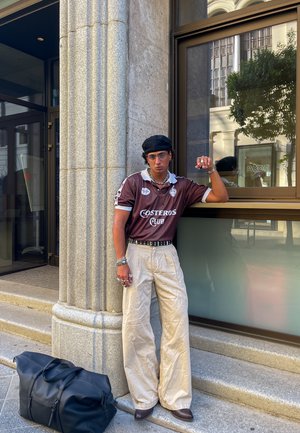 Camiseta de fútbol marrón con acentos blancos, pantalones de pierna ancha beige, boina negra, joyería de plata, de pie cerca de una ventana de cristal, bolsa de deporte negra cerca.