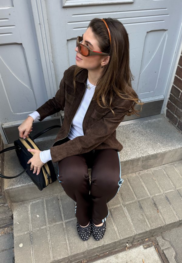 Mujer con cabello castaño y gafas de sol sentada en un escalón de piedra, lleva chaqueta marrón, pantalones oscuros con rayas blancas, zapatos de lunares, sosteniendo una bolsa negra.