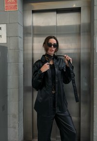 Μαύρο πουκάμισο από τεχνητό δέρμα με κουμπιά και γιακά, συνδυασμένο με μαύρο παντελόνι. Το μοντέλο φοράει ένα μαύρο τοπ από δαντέλα και γυαλιά ηλίου, κρατώντας έναν αντίστοιχο ιμάντα.