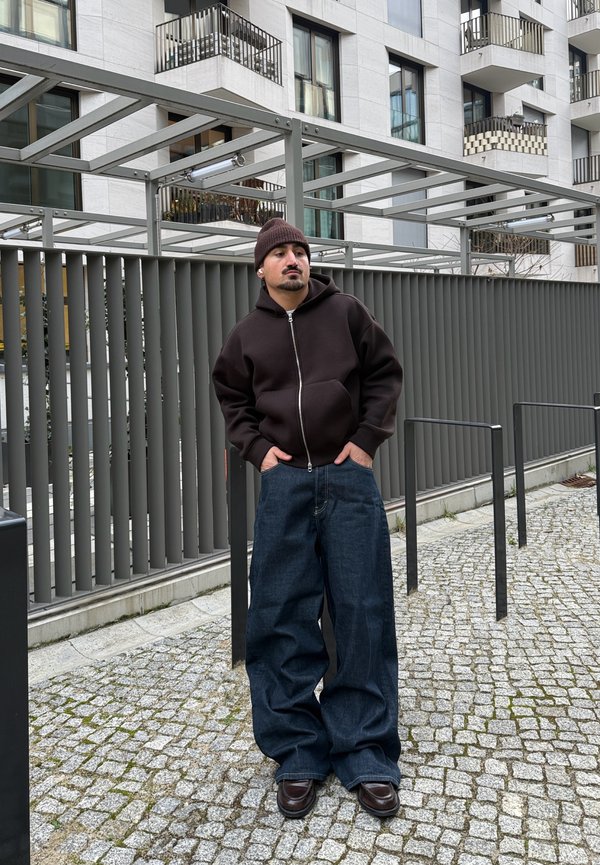 Mężczyzna w ciemnej bluzie z kapturem, czapce typu beanie i szerokich dżinsach stojący na brukowanym chodniku w pobliżu nowoczesnego budynku z metalowym ogrodzeniem.