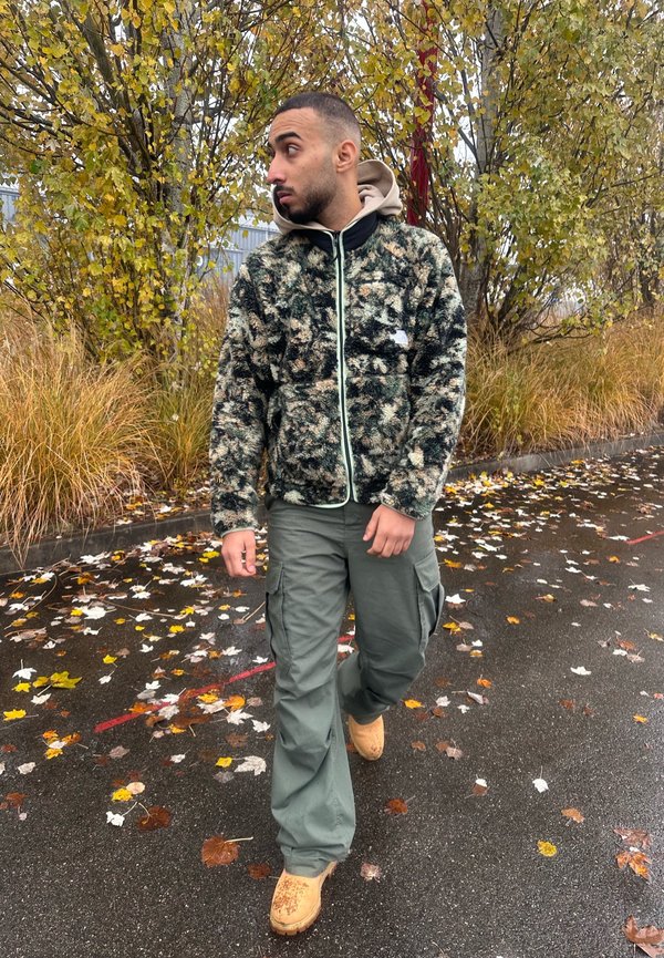 Flauschige grüne Tarnjacke mit schwarzem Saum, grauer Kapuzenpulli darunter, cargo-grüne Hose und sandfarbene Arbeitsstiefel, die auf einem mit Blättern bedeckten Weg gehen.