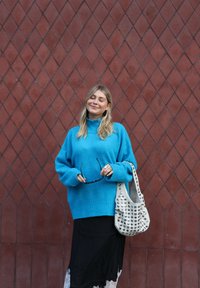 Obszerny turkusowy sweter z dzianiny z wysokim kołnierzem, zestawiony z czarną spódnicą ozdobioną koronką oraz białą torebką z ćwiekami.