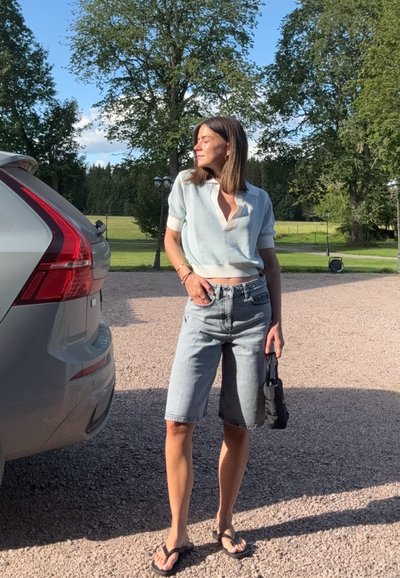 Camiseta recortada de malla azul claro con escote en forma de V, combinada con pantalones cortos de denim claro. Un bolso negro y chanclas complementan el conjunto.
