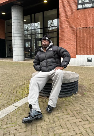 Chaqueta de plumas negra, sudadera gris, pantalones de chándal grises y botas negras brillantes. Sentado en un banco de piedra circular, en un entorno urbano con fondo de pared de ladrillo.