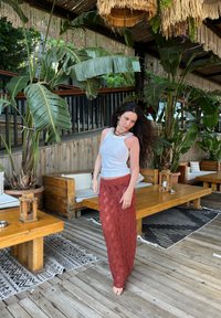 Žena u uskoj bijeloj majici i dugoj, teksturiranoj crvenoj suknji stoji na drvenom podu s velikim zelenim biljkama u pozadini.