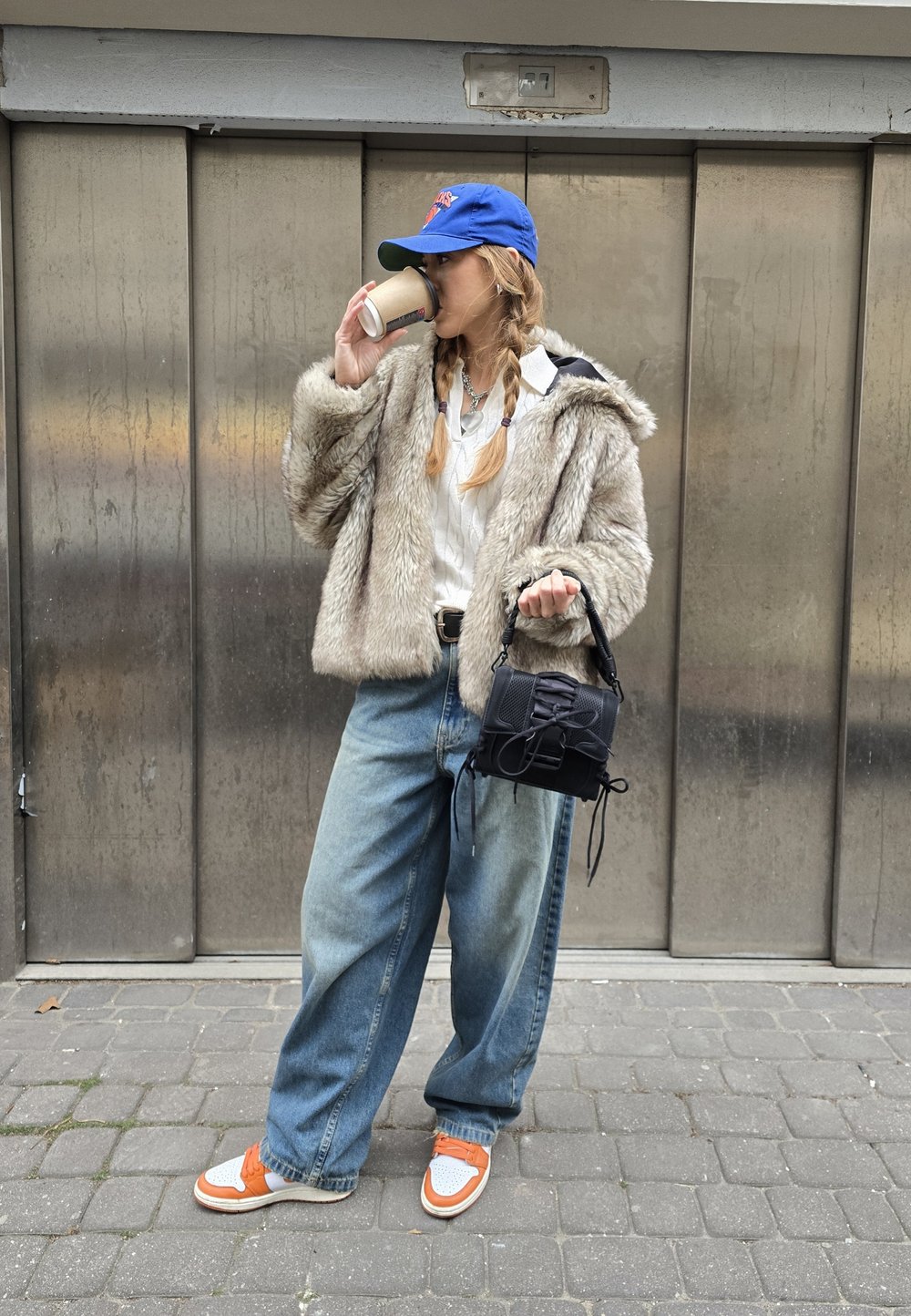 Faux-Fur-Mantel in hellem Beige, darüber ein weißes Hemd, übergroße blaue Jeans, Sneakers mit orangefarbenen Akzenten und eine kleine schwarze Handtasche.