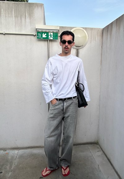 Hombre con gafas de sol, camisa blanca de manga larga, vaqueros grises, sandalias rojas y sosteniendo una bolsa negra, de pie junto a una pared con un cartel de salida de emergencia.