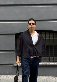 Mann mit Sonnenbrille, schwarzem offenem Hemd über weißem Tanktop, dunklen Jeans, der eine schwarze Tasche hält und vor einer grauen Wand mit vergittertem Fenster steht.