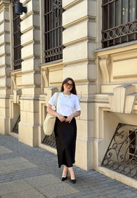 Weißes T-Shirt, schwarze Midirock und schwarze spitze Absätze. Accessoire: geflochtene Tasche. Hintergrund zeigt ein Steinbau mit dekorativen Fenstern.