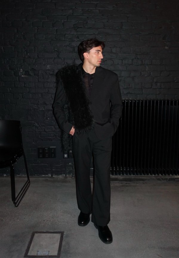 Zwart tailleur met een glanzende afwerking, met een dikke nepbontaccent op één schouder. Matte zwarte schoenen maken de outfit compleet.