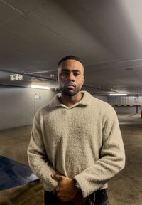 Homme aux cheveux courts et barbe, portant un pull crème texturé, debout les mains croisées dans un parking couvert.