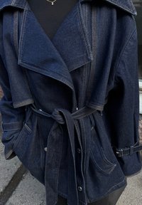 Temno modra jeans jakna s širokim ovratnikom, pasom in dvema sprednjima žepoma. Ponuja kontrastno šivanje in manšete z nastavljivimi trakovi.