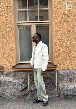 Persona con una chaqueta crema sobre una camisa estampada, pantalones vaqueros claros y calzado negro, de pie junto a una pared naranja texturizada y una ventana.