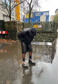 Svarta vattentäta stövlar med ljusa snören och kontrasterande sula, kombinerade med svarta lädershorts och en matchande jacka, stående i grunt vatten.